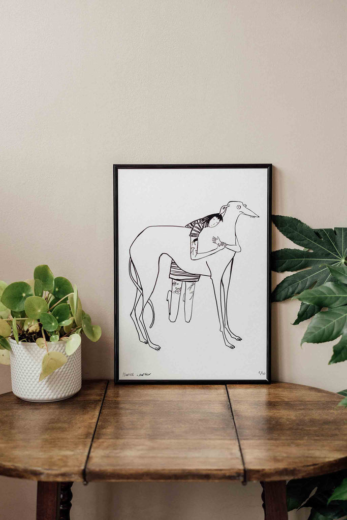 Black line drawing of tattooed girl in stripy dress hugging large derpy greyhound/lurcher/sighthound on white background print in black frame resting on table